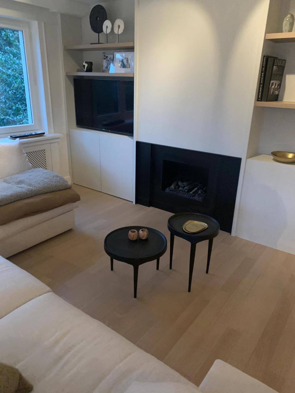 Ingerichte woonkamer met een zeer lichte eiken vloer, zwarte bijzettafels en een zwarte haard